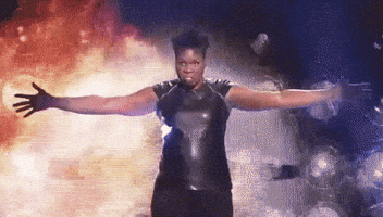 SNL gif. Leslie Jones, dressed in a shiny black top, stands in front of what looks like an explosion with her arms spread wide, threateningly staring ahead, and then dropping her arms to her side.