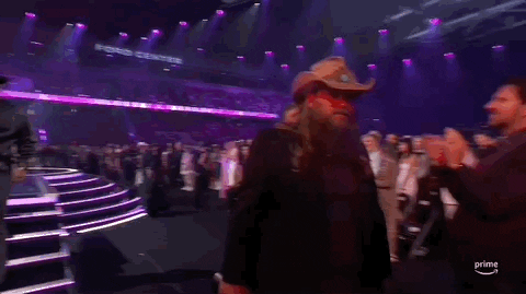 Academy of Country Music Awards gif. Chris Stapleton walks past standing audience members with a pleased look on his face, nods his head to acknowledge a couple of people while nondescriptly saying something.