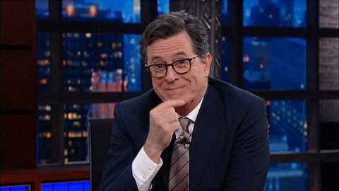 Celebrity gif. Stephen Colbert on The Late Show leaning onto his desk and suppressing a laugh with his hand in front of his mouth.