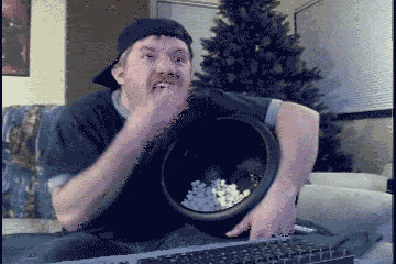 Video gif. Man with a backwards hat sits on the edge of a sofa with a bucket of popcorn on his lap, his eyes glued to whatever’s in front of him. He mindlessly grabs handfuls of popcorn and stuffs them in his mouth. He chomps down, not caring that kernels fall out of his mouth.
