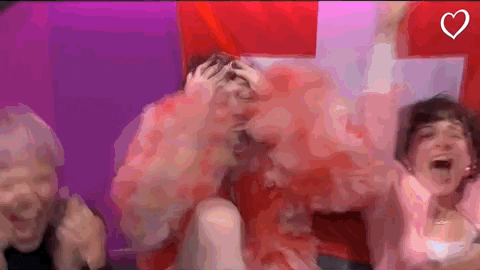 TV gif. Swiss musician Nemo is dressed in a pink skirt and chiffon pink top at the 2024 Eurovison Song Contest. They grasp their head and excitedly gasp and scream as they hug the person next to them. The flag of Switzerland can be seen in the background.
