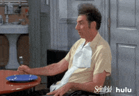 Seinfeld gif. Michael Richards as Kramer gazes forward at a table and idly taps his foot and his fist in time, holding a fork.