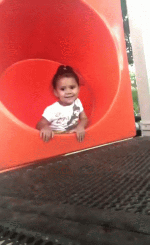 Video gif. A little girl peeks out of an orange tube slide and waves before disappearing down the slide backwards and belly down with her hands in the air. 