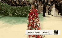 Met Gala 2024 gif. Jessica Serfaty Michel wearing a red Dolce and Gabbana cape covered in large 3D flowers, arrives and adjusts her ensemble to reveal a plunging halter bodice.
