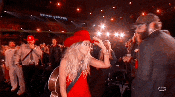 TV gif.  Country music artist Lainey Wilson walking through the crowd of excited fans at a live concert event. She is wearing a vibrant red dress and a matching cowboy hat. She engages with an audience member, placing her red cowboy hat on the woman’s head.