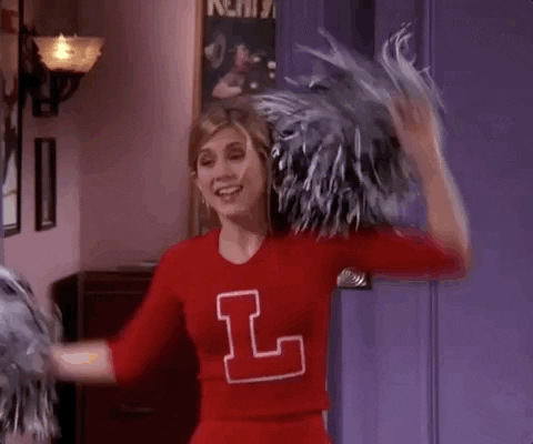 Friends gif. Dressed up as a cheerleader, Jennifer Aniston as Rachel holds up her pom poms and jumps in excitement.