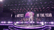 Academy of Country Music Awards gif. Chris Stapleton walks on stage and shakes the hands of the award presenters awaiting him. The words in the background read "Male Artists of the Year". As one presenter hands Stapleton the throphy, the other walks off the stage as if his job his done. Chris holds appreciative demeanor.