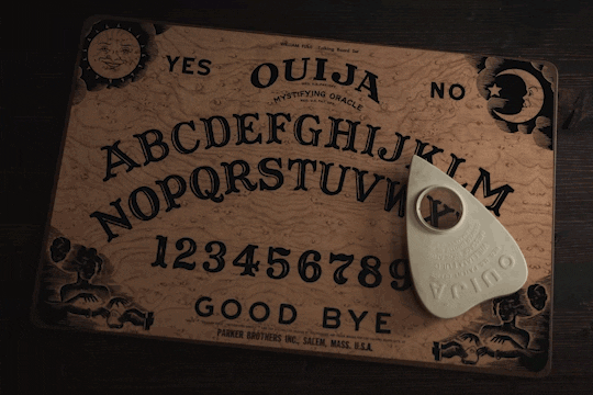 Video gif. A wooden ouija board lays on a table. The corners of the ouija board have ornate, black and white illustrations of the sun, the moon, and people using the ouija board. The board has the whole alphabet, numbers zero to nine, goodbye written at the bottom, and yes and no written at the top. The heart shaped planchette moves across the board from the letter X to the word “yes” all on its own. 
