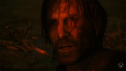 Video game gif. Close-up of a man with long disheveled hair and a beard in the game "Alan Wake." He mumbles or chants softly, looking off towards a red glowing light that illuminates his face. 