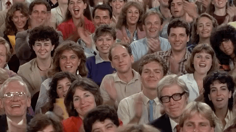 Video gif. Crowd of people who look like they’re from the 1980s looks at us and claps all in unison, and smile.