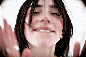 Music video gif. A scene from Billie Eilish's music video for "LUNCH" shows a close-up of Billie's face—who has long black hair—against a stark white background taken from a fisheye lens. She playfully bites her bottom lip and smiles while clapping for the camera.