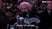 Movie gif. Christopher Walken as Max in Batman Returns stands at a microphone making a speech with a large outdoor Christmas tree behind him, as he waves and says, "Merry Christmas."