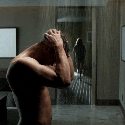 Movie gif. Jamie Dornan as Christian Grey in "Fifty Shades of Grey" stands in the shower, running his hands through his hair, as Anastasia, played by Dakota Johnson, walks toward him from the background.