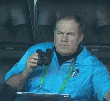 Sports gif. Bill Belichick grabs a snack from his seat in the stands, then peers intently through binoculars as he munches.