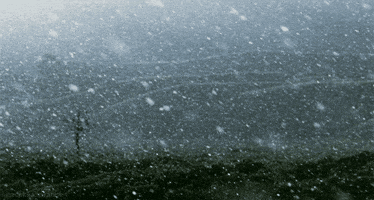 Video gif. Snow falls heavily on a hilly landscape.