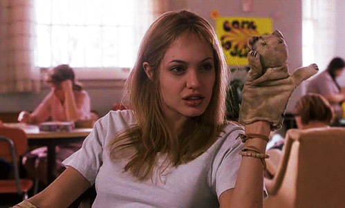 Movie gif. Angelina Jolie as Lisa Rowe from Girl, Interrupted sits wearing a cat hand puppet, which moves like it's waving at someone.