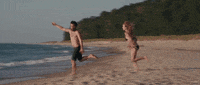Movie gif. On a beach, Olivia Wilde as Kate in Drinking Buddies gallops after Jake Johnson as Kyle, who trips and falls once he reaches the water.