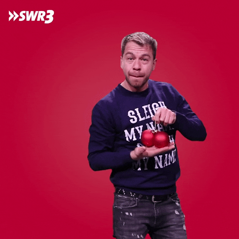 Video gif. Kemal Goga from the German radio show SWR3, holds up two red ball ornaments and cups them in his other hand.