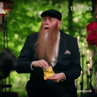 Sponsored gif. Bearded man in a black suit and cap eats chips from a bag while watching something with a look of intense interest. In the upper right corner is the logo for the show The Traitors.