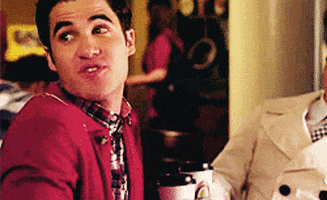 Celebrity gif. Darren Criss looks over his shoulders at someone and smiles while crossing his fingers, wishing them luck.