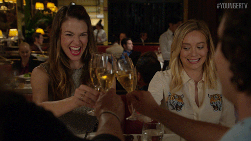 TV gif. Sutton Foster as Liza and Hilary Duff as Kelsey in Younger. The two are on a double date and everyone clinks wineglasses as Kelsey looks down shyly and Liza smiles broadly.
