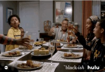 TV gif. The Johnson family in black-ish are rising from their seats at the dinner table to toast during their Thanksgiving meal.