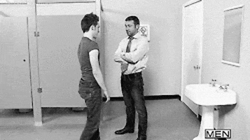 Video gif. Two men are in a public bathroom. One man with a tie stands with his arms crossed as a man in a t-shirt walks up to him. The man with the t-shirt as he holds onto the other man's hips then drops into a full split. The man with the tie looks surprised and impressed.