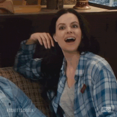 TV gif. Emily Hampshire as Stevie Budd on Schitt's Creek sits in a chair, relaxed. She looks at someone with a sarcastic enthusiastic look on her face, pretending to be unbothered, and says, “Oh hi!”