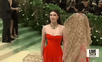 Met Gala 2024 gif. Closeup of Nell Diamond from above smiling for pictures wearing a cherry red Hill House Home sleeveless gown with a gold and diamond necklace, pink blush, and long straight hair with a structured bump at the top of her head.