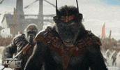 Trailer gif. A daytime scene from the movie "Kingdom of the Planet of the Apes" shows an intimidating Proximus Caesar wearing a three-horned metal crown as he smirks and lurches forward in a show of dominance. An imposing gorilla with a taser spear can be seen behind him as a grunt.