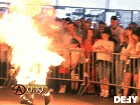 TV gif. Criss Angel walks around with his entire back on fire, a large crowd spectating him from behind metal barricades. A gold timer with white text in the bottom left counts from nineteen seconds to twenty-four seconds.