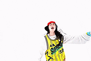 Music Video gif. From Billie Eilish's music video for "LUNCH", Billie is on a stark white background dressed in a long sleeve white shirt with a yellow and black jersey over the top, with a red hat on. She is dancing and singing as she gets closer to the camer, and then walks backwards as the camera pans back.
