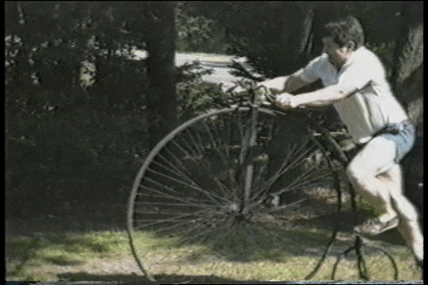 Video gif. A man gets up on a vintage bicycle where the front wheel is super big and the back is small. After hopping on, the bicycle rolls forward and he tips off , falling down on the ground. 
