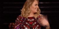 Celebrity gif. Adele turns to the side and erupts in a surprised smile as she waves off camera.
