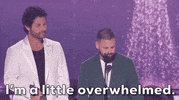 TV gif. Dan Smyers and Shay Mooney accepting an award at the 2024 ACM Awards, both wearing a suit jacket over a t-shirt. Shay is speaking into the microphone. The text overlay at the bottom reads,  'I'm a little overwhelmed.'