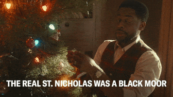 TV gif. Dule Hill, as Bill in The Wonder Years places an ornament onto a lit Christmas tree as he says, “The real St. Nicholas was a Black Moor.”