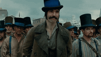 Movie gif. Daniel Day Lewis as Bill the Butcher in Gangs of New York stands menacingly in front of his top hat wearing street gang while they help remove his coat before battle. 
