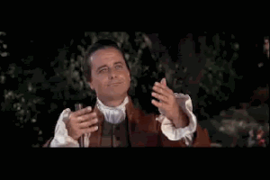 Video gif. Man in old colonial clothes gestures his hands in front of him to present something with a sarcastic smile. He then looks away with an irritated look on his face.