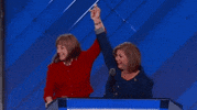 Politics gif. California representatives Loretta Sanchez and Linda Sanchez, wearing a red and a blue outfit respectively, hold hands, waving them in the air at the DNC 2016 podium.