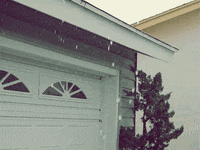 Video gif. Hail falls in front of a white garage that has a lonely tree resting next to it.