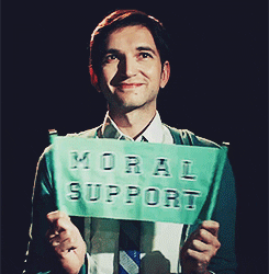 Video gif. Man looks up with a warm smile as he holds a small banner that says, “Moral support.” He shakes the banner a bit to give it emphasis.