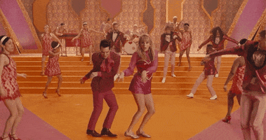 Music video gif. From Taylor Swift's Me, Taylor and Brendon Urie dance The Twist with their hands held up like paws, bent at the wrist. They are at the center of a bright orange and pink 60s-style stage set with about a dozen dancers filling the background. 