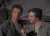 TV gif. Alan Alda as Capt. Benjamin and Wayne Rogers as Capt. John in MASH. They both hold up cocktail glasses and point at someone before moving towards them to cheers together.