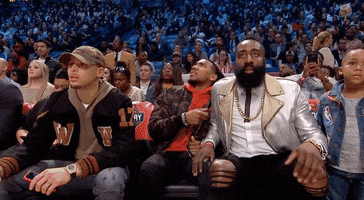Celebrity gif. James Harden leaps from his courtside chair as he yells and claps excitedly. 