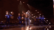 ACM Awards gif. Post Malone taps heel to the beat of the song with a guitar strapped around him as he sings into the standing mic while holding it with both hands and briefly shaking his head from left to right. 