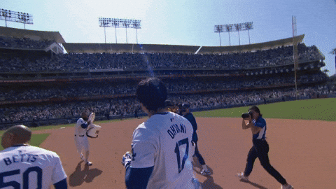 Sports gif. Shohei Ohtani of the Los Angeles Dodgers is doused with a bucket of water and he runs around the field with his teammates.