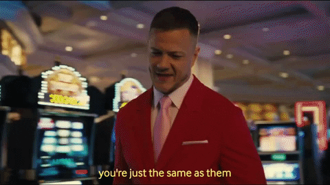 Music video gif. Imagine Dragons frontman Dan Reynolds walks through a casino and reaches into the pocket of his red sport jacket. Text, "You're just the same as them."