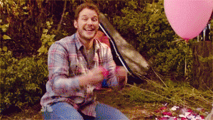 Parks and Recreation gif. Chris Pratt as Andy sits on a picnic blanket in the woods, picking up pieces of red, white, and pink colored paper and throwing them dramatically in the air over his head.