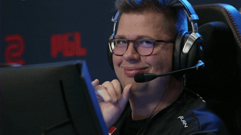Sports gif. FaZe Karrigan sits in a chair with headphones on at an esports event and waves at us, wiggling his eyebrows and blowing us a kiss as we zoom in.