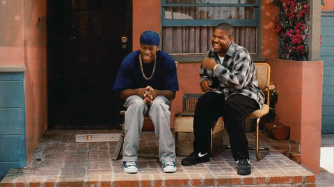 Celebrity gif. Ice Cube and Chris Tucker are sitting on a patio and they're both laughing heartily. Ice Cube puts his hand over his face while laughing and Chris claps while lifting both his feet up.
